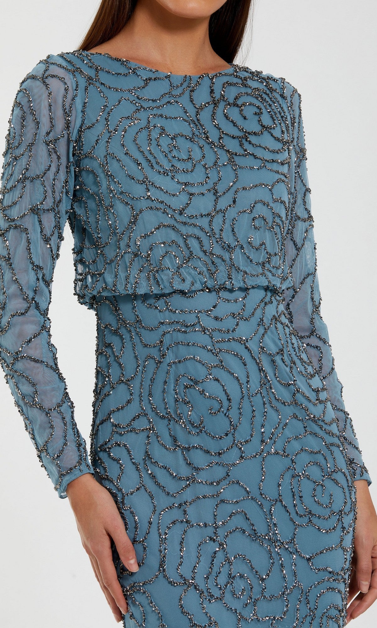 A woman models the Mac Duggal 6189 Long Formal Dress, a fitted, long-sleeve blue gown adorned with an elegant rose beaded pattern. The image captures her from shoulders to mid-thigh, with one arm bent and the other resting at her side.